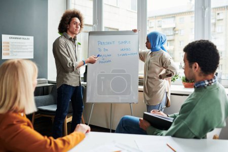 Photo for Young confident student or teacher making presentation of Present Simple tense while standing by whiteboard in front of audience - Royalty Free Image