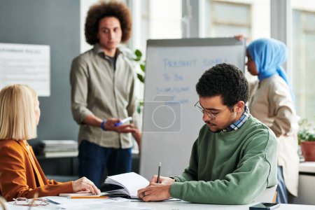 Young man making notes in copybook while sitting by desk at lesson of English grammar against guy explaining new subject to women