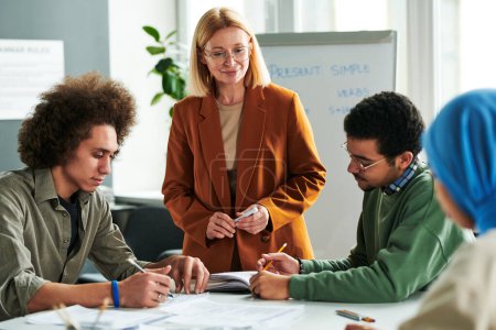 Photo for Confident teacher standing in front of group of young intercultural students making notes in their copybooks at lesson or seminar - Royalty Free Image