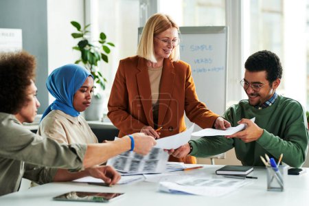 Photo for Happy mature teacher passing papers with grammar tests to group of multicultural students sitting by table at lesson of English language - Royalty Free Image