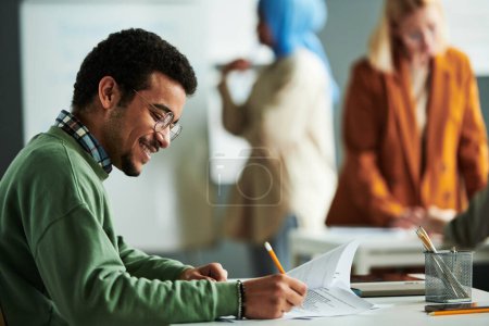 Photo for Side view of happy Middle Eastern student putting ticks in front of right answers while carrying out grammar test by desk in classroom - Royalty Free Image