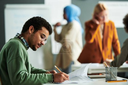 Photo for Young serious student in eyeglasses and casual pullover making notes on paper with exam text during individual work at lesson - Royalty Free Image