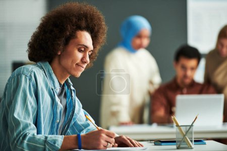 Foto de Vista lateral de un joven estudiante multiétnico mirando papel con prueba de gramática y señalándolo mientras piensa en la respuesta correcta en la lección - Imagen libre de derechos