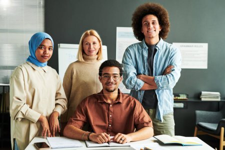 Photo for Group of multicultural students and mature blond teacher in casual attire standing by desk in front of camera with whiteboard behind - Royalty Free Image