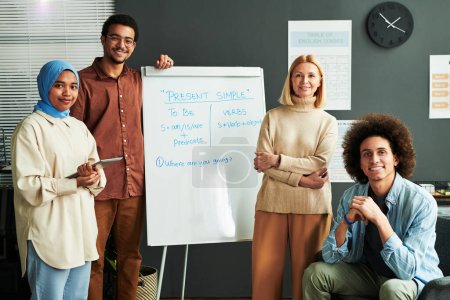 Photo for Several happy young intercultural students and mature blond teacher surrounding whiteboard with explanation of Present Simple tense - Royalty Free Image