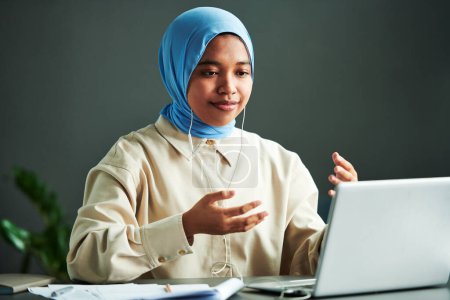 Téléchargez les photos : Jeune femme musulmane confiante en tenue décontractée et hijab bleu expliquant quelque chose alors qu'elle était assise devant un ordinateur portable pendant la leçon en ligne - en image libre de droit