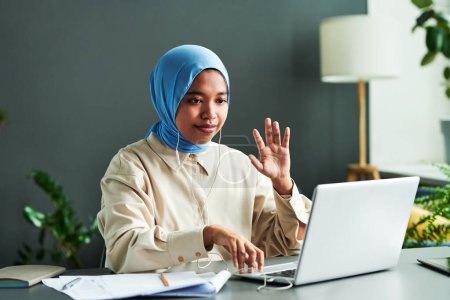 Photo for Young Muslim student in blue hijab greeting teacher by waving hand during online lesson while sitting by desk in front of laptop - Royalty Free Image
