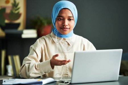 Téléchargez les photos : Jeune femme musulmane confiante dans le hijab prenant part à un webinaire assis à côté du bureau devant un ordinateur portable à la maison et expliquant quelque chose - en image libre de droit