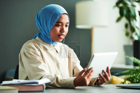 Téléchargez les photos : Femme musulmane confiante enseignant ou étudiant en hijab bleu regardant le public en ligne sur l'écran de la tablette tout en communiquant avec eux - en image libre de droit