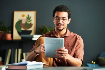 Junge männliche Studenten aus dem Nahen Osten betrachten Online-Informationen auf dem Bildschirm eines Tablets, während sie mit Büchern am Arbeitsplatz sitzen