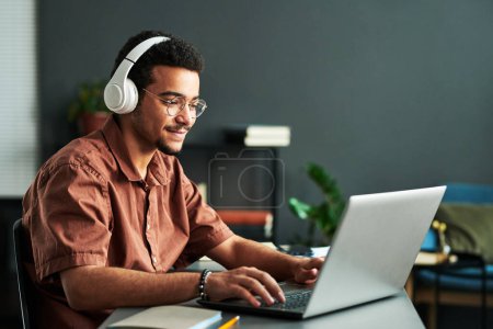 Junger lächelnder Mann mit Kopfhörer tippt auf der Laptop-Tastatur, während er am Arbeitsplatz sitzt und an einem Online-Webinar oder Unterricht teilnimmt