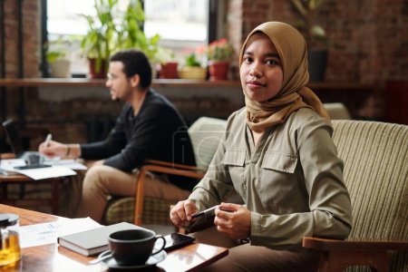 Foto de Joven mujer de negocios bonita en hijab sentada junto a la mesa frente a la cámara y mirándote mientras organizas el trabajo contra tu colega - Imagen libre de derechos