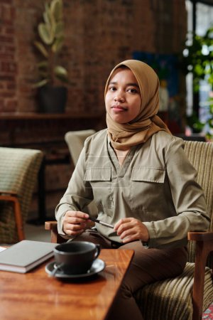 Téléchargez les photos : Jeune employée musulmane afro-américaine réussie ou stagiaire en hijab et vêtements décontractés assis dans un fauteuil et organisant le travail - en image libre de droit