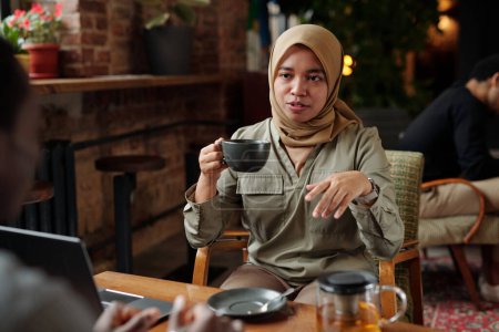 Foto de Joven mujer de negocios musulmana en hijab tomando té y hablando con un colega masculino en la reunión de trabajo mientras está sentada en sillón por mesa - Imagen libre de derechos