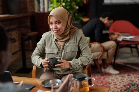 Téléchargez les photos : Jeune employée musulmane dans le hijab tenant une tasse de tisane alors qu'elle était assise à table devant un collègue masculin pendant la conversation - en image libre de droit