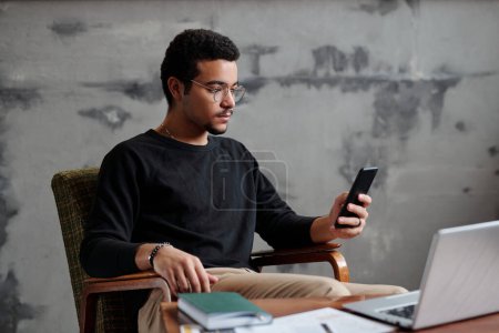 Foto de Joven hombre de negocios serio mirando la pantalla del teléfono inteligente mientras está sentado en el sillón por el lugar de trabajo contra la pared gris en la oficina y mensajes de texto - Imagen libre de derechos