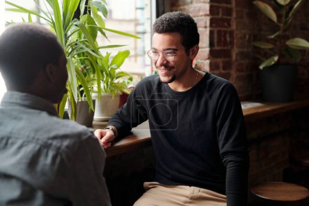 Téléchargez les photos : Jeune homme d'affaires souriant en tenue décontractée regardant un collègue afro-américain avec le sourire lors de la discussion des points d'organisation - en image libre de droit