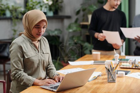 Téléchargez les photos : Jeune femme d'affaires sérieuse dans le hijab regardant l'écran d'ordinateur portable tout en étant assis par le lieu de travail et en organisant le travail contre des collègues - en image libre de droit