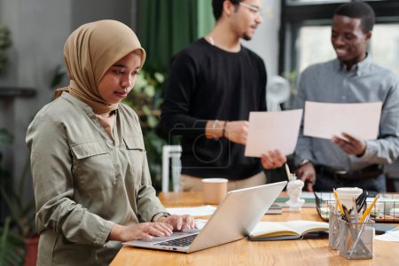 Téléchargez les photos : Jeune femme d'affaires musulmane organisant le travail devant un ordinateur portable tout en se tenant debout sur le lieu de travail contre deux hommes interculturels avec des papiers - en image libre de droit