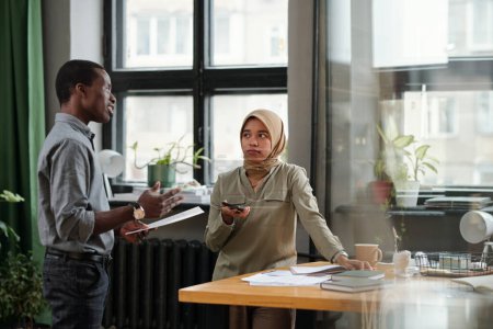 Photo for Young Muslim businesswoman listening to confident African American male colleague with document making report at meeting - Royalty Free Image