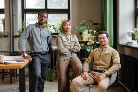 Téléchargez les photos : Portrait de trois jeunes collègues interculturels en tenue décontractée intelligente regardant la caméra tandis que le leader confiant de l'équipe d'affaires assis dans un fauteuil - en image libre de droit