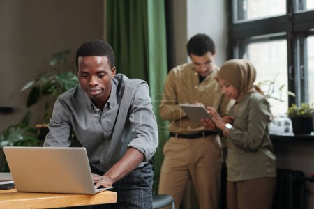 Foto de Joven hombre de negocios afroamericano inclinado sobre el escritorio con computadora portátil y la creación de redes contra dos colegas interculturales utilizando tableta - Imagen libre de derechos