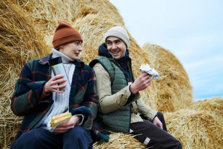 Téléchargez les photos : Deux jeunes travailleurs heureux de la ferme en vêtements de travail assis par pile de foin devant la caméra, parlant et ayant des sandwichs et du thé pour le déjeuner - en image libre de droit