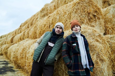 Photo for Two young confident workers of modern farm in casualwear looking at camera while standing by huge stack of fresh hay in the country - Royalty Free Image
