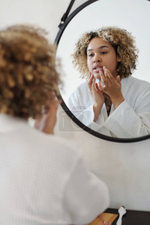 Foto de Joven mujer afroamericana mirando en el espejo mientras se aplica crema hidratante en la cara durante la rutina de la mañana - Imagen libre de derechos