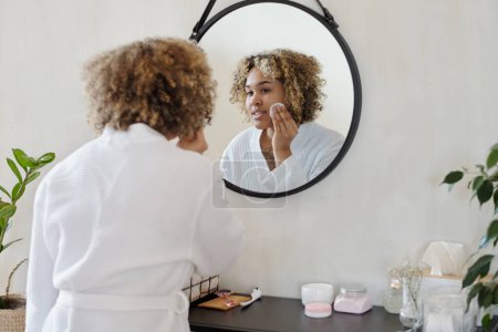 Téléchargez les photos : Jeune jolie femme en peignoir blanc appliquant tonifiant rafraîchissant sur le visage avec du coton tout en se tenant devant le miroir à la maison - en image libre de droit