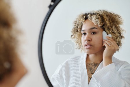 Photo for Young woman in bathrobe massaging her face with gua sha massager after morning hygiene procedures while standing in front of mirror - Royalty Free Image