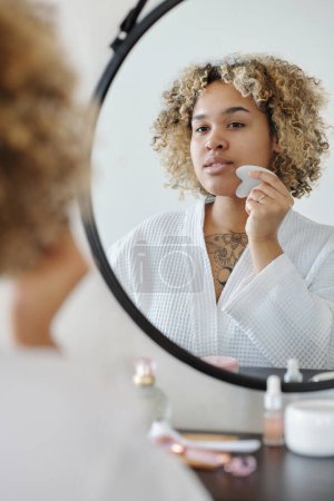 Foto de Joven mujer afroamericana con rascador de cuarzo masajeando su cara frente al espejo en el baño después de los procedimientos de cuidado de la belleza matutina - Imagen libre de derechos
