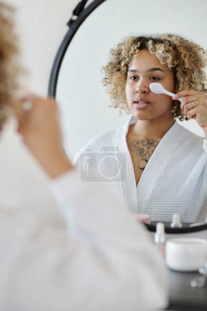 Téléchargez les photos : Jeune femme utilisant l'outil de traitement beautycare tout en se tenant devant le miroir dans la salle de bain et en prenant soin de son visage le matin - en image libre de droit