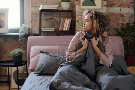 Téléchargez les photos : Jeune femme noire détendue en pyjama couvrant de couverture grise alors qu'elle était assise sur le lit le matin après avoir dormi réticente à se lever - en image libre de droit