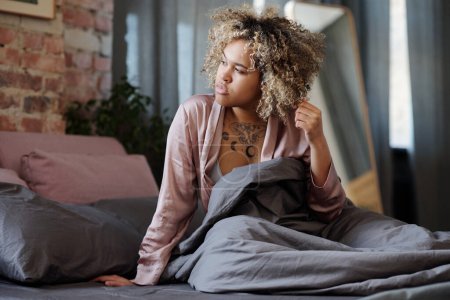 Foto de Mujer joven perezosa en pijama de seda mirando a través de la ventana del dormitorio mientras se sienta en la cama bajo una manta gris después de despertarse por la mañana - Imagen libre de derechos
