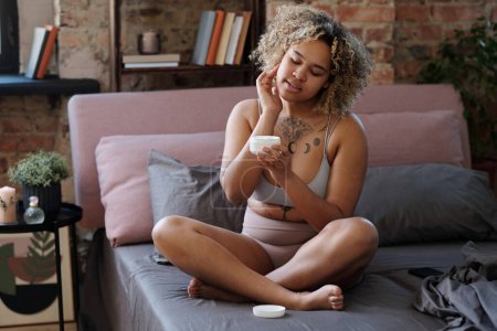 Téléchargez les photos : Jeune femme afro-américaine en sous-vêtements avec pot de crème l'appliquer sur son visage tout en étant assis sur un lit double le matin dans la chambre - en image libre de droit