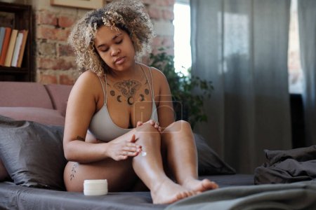 Téléchargez les photos : Jeune femme afro-américaine nourrissant la peau avec de la lotion corporelle tout en étant assis sur le lit le matin après avoir pris une douche et se raser les jambes - en image libre de droit