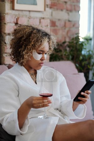Foto de Joven mujer afroamericana con parches bajo los ojos usando un teléfono inteligente y tomando un poco de vino tinto mientras se relaja en la cama por la mañana - Imagen libre de derechos