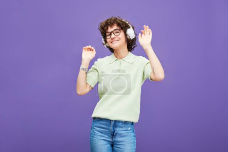 Téléchargez les photos : Jeune femme joyeuse en casque et vêtements décontractés écouter de la musique et danser devant la caméra sur fond de lavande en isolement - en image libre de droit