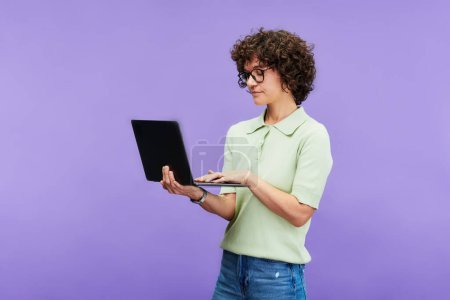 Téléchargez les photos : Jeune femme brune sérieuse en tenue décontractée tapant sur clavier d'ordinateur portable tout en réseautant sur l'étude en ligne sur fond lilas - en image libre de droit