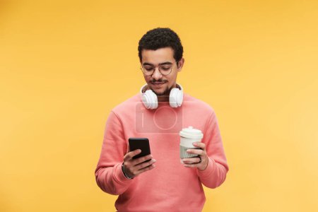 Téléchargez les photos : Jeune homme multi-ethnique avec casque blanc autour du cou à l'aide d'un smartphone et ayant une tasse de café sur fond jaune - en image libre de droit