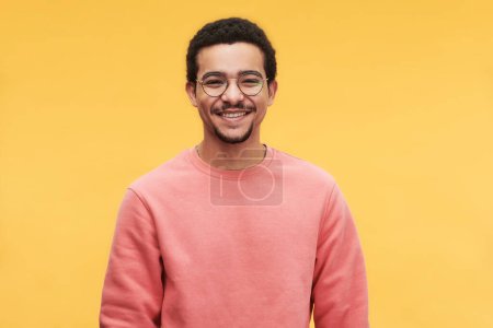 Téléchargez les photos : Jeune homme joyeux en lunettes et sweat-shirt rose regardant la caméra tout en se tenant debout sur un fond jaune vif en isolement - en image libre de droit