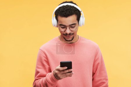 Téléchargez les photos : Jeune homme en pull rose écoutant de la musique dans les écouteurs et choisissant de nouvelles pistes sonores dans le téléphone mobile devant la caméra - en image libre de droit