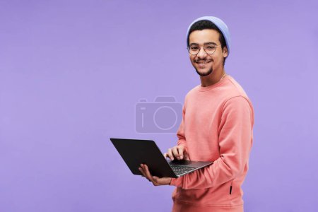 Photo for Young successful designer or freelancer in casualwear using laptop and looking at camera with smile while standing on violet background - Royalty Free Image