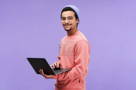 Foto de Joven y exitoso freelancer masculino o programador en jersey rosa, gorro de gorro y anteojos sosteniendo laptop y mirando la cámara - Imagen libre de derechos