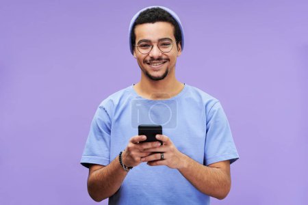 Téléchargez les photos : Jeune homme souriant en tenue décontractée bleue regardant la caméra tout en envoyant des SMS dans le smartphone ou en faisant défiler les photos sur fond violet - en image libre de droit