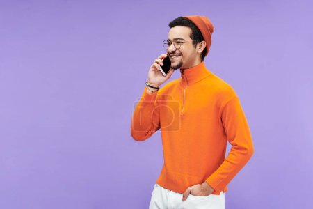 Téléchargez les photos : Joyeux jeune homme au col roulé, bonnet et lunettes communiquant sur smartphone tout en se tenant devant la caméra - en image libre de droit
