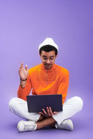 Téléchargez les photos : Jeune homme regardant l'écran d'ordinateur portable avec étonnement tout en étant assis devant la caméra avec ses jambes croisées et en réseautage - en image libre de droit