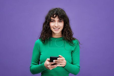 Photo for Young woman with dark long wavy hair looking at camera and using mobile phone while standing in isolation on violet background - Royalty Free Image