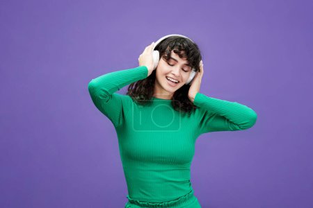 Foto de Joven morena alegre con el pelo largo y ondulado escuchando música en los auriculares y disfrutando mientras mantiene los ojos cerrados - Imagen libre de derechos